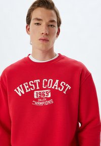 Sudadera roja con estampado en blanco "CAMPEONES DE LA UNIVERSIDAD DE LA COSTA OESTE 1965", cuello redondo y corte relajado. Textura suave con cuello de canalé.