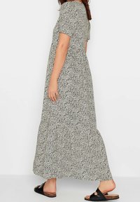 Maxi sukienka z białej tkaniny w czarne plamki, z krótkimi rękawami i warstwowym falbanowym dołem, noszona z czarnymi sandałami i długości odsłaniającej kostki.