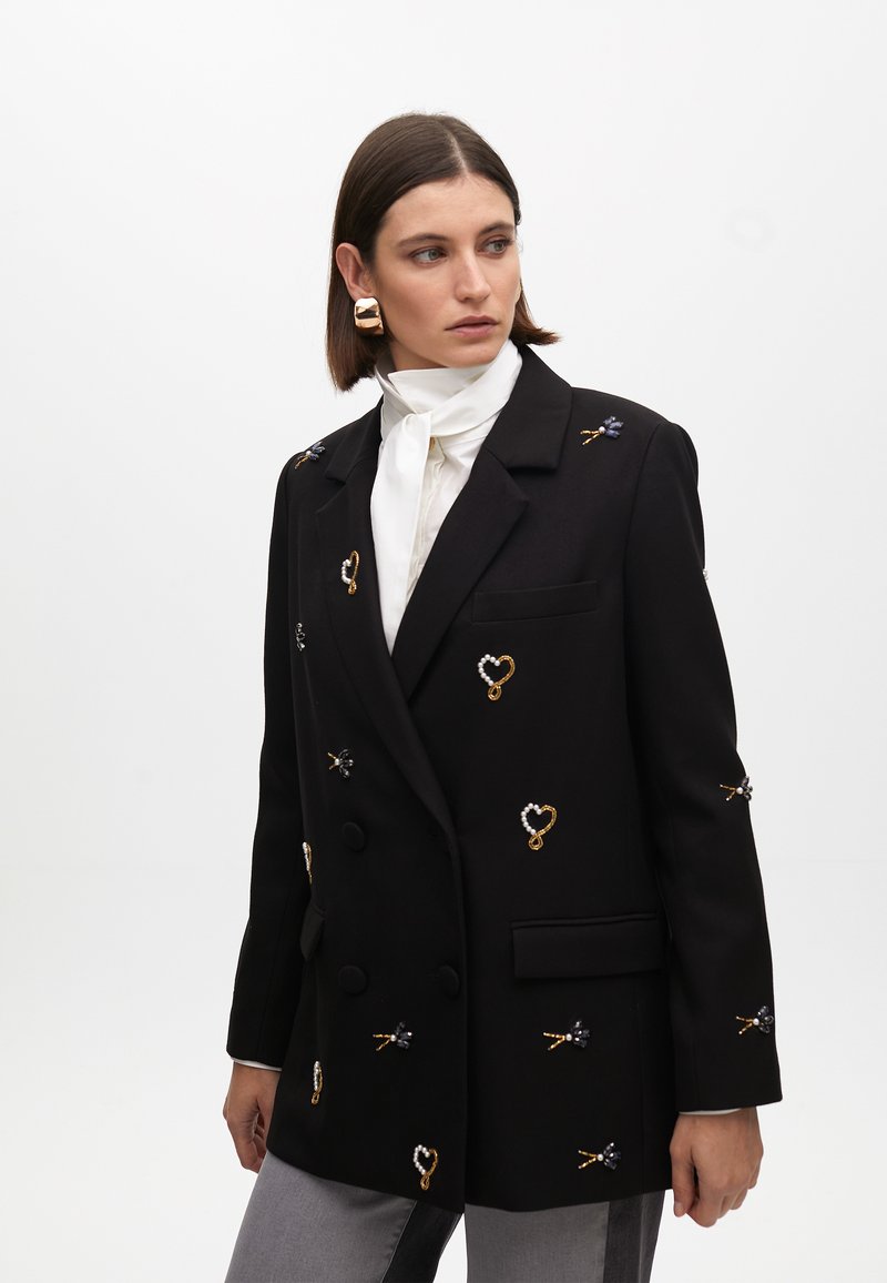 Blazer nero doppiopetto con decorazioni in perline e ricami in oro e blu. Presenta un colletto a punta e tasche frontali.