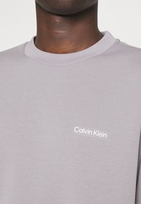Sweatshirt grigio chiaro a girocollo realizzato in tessuto morbido, con un piccolo logo bianco "Calvin Klein" sul lato sinistro del petto.