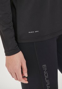 Musta urheilullinen pitkähihainen paita, jossa valkoisella painettuna "QUICK DRY"; istuva malli sileällä tekstuurilla, yhdistettynä mustiin leggingsseihin, joissa on "ENDURA" -brändäys.