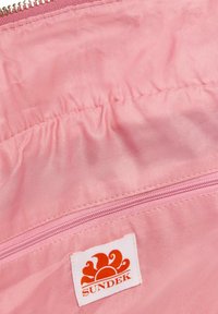 Tissu en nylon rose avec un design froncé, comportant une fermeture éclair et une étiquette blanche avec un logo de soleil orange portant l'inscription "SUNDEK".