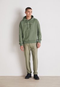 Felpa verde in cotone con tasca anteriore e cordini, abbinata a pantaloni grigi chiari. Sneakers nere completano l'outfit.