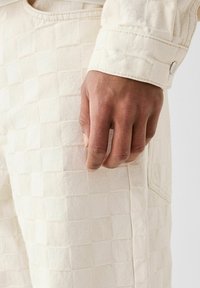 Krämfärgade texturerade byxor med rutmönster och en sidficka, med en knappstängning vid fållen och en slät yta.