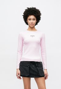 Top de manga comprida rosa claro com textura canelada e decote redondo, com o logótipo preto "GUESS". Combinado com uma saia de ganga preta com bolsos.