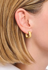 Goldfarbene Creolen mit einem leicht gebogenen, polierten Design, die am Ohrläppchen befestigt sind und eine glatte Oberfläche sowie einen modernen Stil aufweisen.