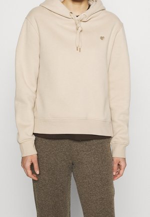 Personne portant un sweat à capuche beige avec des cordons noués et un petit emblème en forme de cœur, associée à un pantalon marron texturé, visible du cou aux hanches.