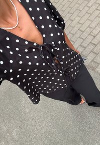Person som bär en svart topp med prickar och knytning fram, svarta byxor, vit pärlhalsband med stjärnhänge och sandaler, står på asfalten.
