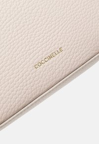 Jasnoróżowa torebka ze skórki teksturowanej o gładkim wykończeniu, z wytłoczonym złotym logo z boku oraz kontrastującym białym wykończeniem.