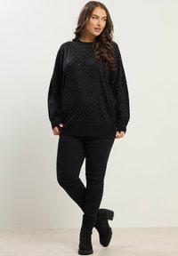 Mujer con cabello largo y ondulado lleva un suéter negro con pequeños puntos blancos, pantalones negros y botas negras hasta el tobillo, de pie contra un fondo liso.