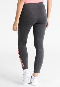 Szare legginsy wykonane z elastycznej tkaniny, o dopasowanym kroju, z wyraźnym różowym logo wzdłuż lewej nogawki. Połączone z białymi butami sportowymi.