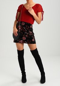 Blouse rouge transparente à manches nouées, associée à une mini-jupe en velours noir à motif floral et des bottes longues en daim noir. Sac bandoulière rond noir.