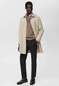 Abrigo ligero beige con cierre de botones, llevado sobre un suéter marrón y pantalones negros, complementado con mocasines negros brillantes.