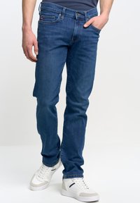 Mees, kes kannab siniseid kitsaid teksapükse ja valgeid tossusid, seisab ühe käega taskus ühevärvilise tausta ees.