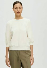 Woman with dark hair in a low bun wearing a white long-sleeve blouse and olive green pants, standing against a plain light background.