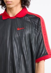 Black Nike sports shirt with red collar, red shoulder stripes, and red Nike swoosh logo on the chest, worn by a person with curly hair.