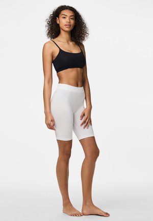 Vrouw met krullend haar, staand op blote voeten, draagt een zwart cropped tanktop en witte hoge taille fietsbroekjes tegen een effen achtergrond.