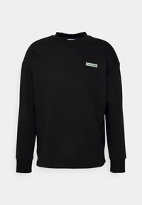 Felpa nera con maniche lunghe e collo a coste, caratterizzata da una piccola patch verde con il logo Calvin Klein sul petto. Texture liscia.