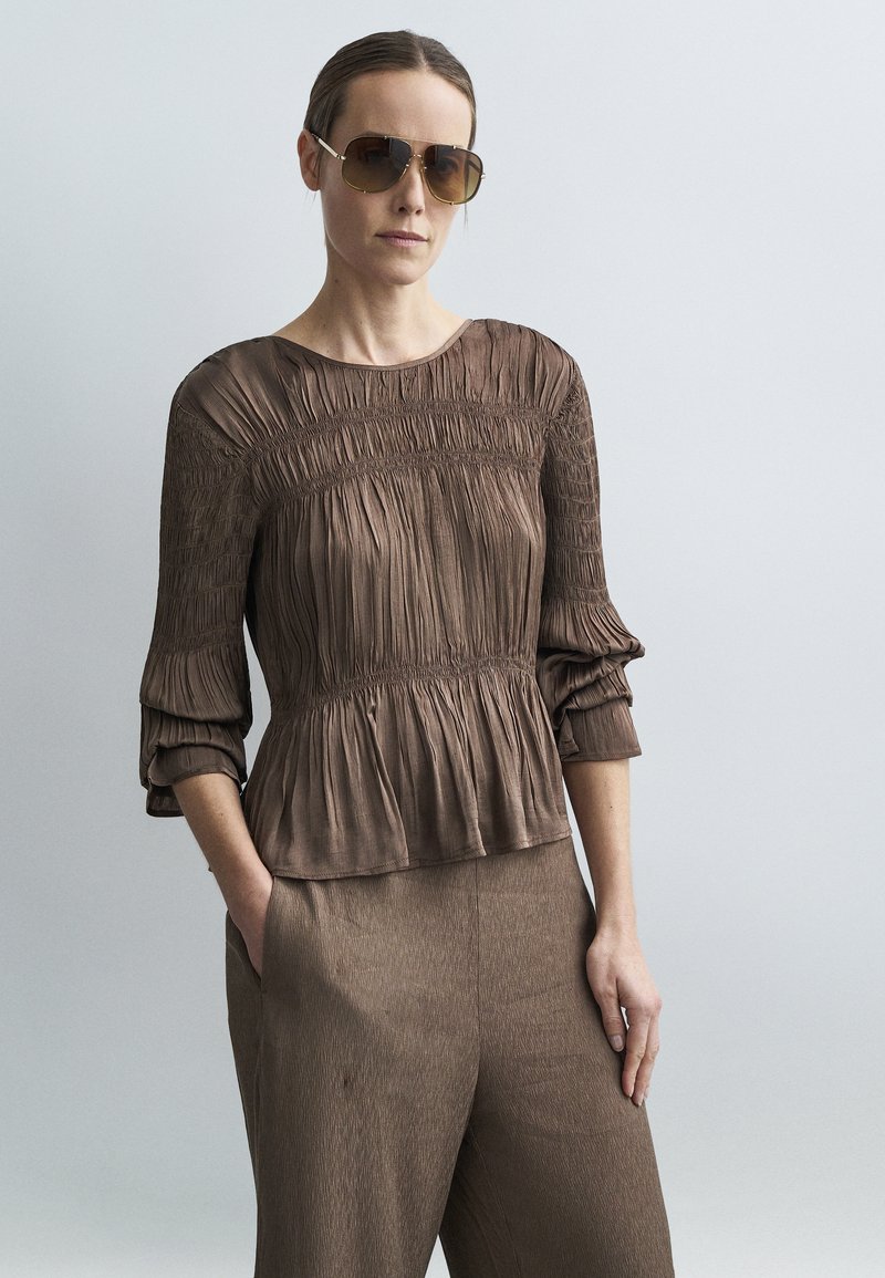 Barna, ráncolt blúz háromnegyedes ujjakkal és húzott derékkal. A modell nagy, színezett napszemüveget visel, amelyhez barna nadrágot párosítottak.