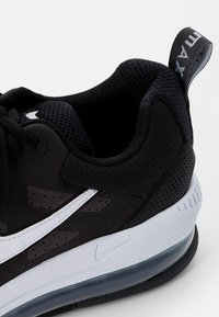 Černá sportovní bota s prodyšnou síťovinou a syntetickými materiály, bílými akcenty a viditelnou vzduchovou jednotkou v podrážce. Černé poutko s textem "Air Max".