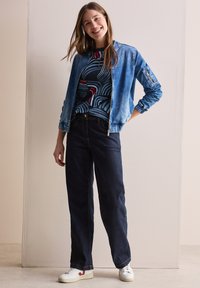 Giacca di denim blu chiaro, con chiusura a zip e polsini a costine, indossata sopra una camicia navy a fantasia. Abbinata a pantaloni di denim scuro e sneakers bianche.