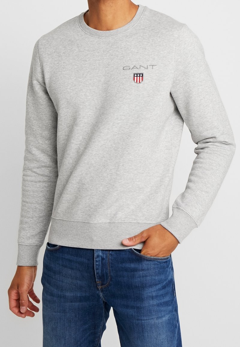 Grijze trui met ronde hals, geribbelde manchetten en zoom. Heeft een geborduurd "GANT" logo en een schild met rood-wit-blauwe accenten.