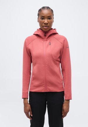 Nő korallrózsaszín cipzáras kapucnis pulóverben és fekete nadrágban, világos szürke egyszínű háttér előtt áll.