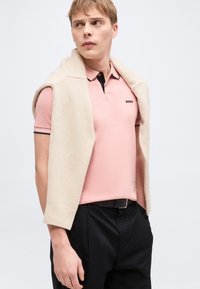 Νέος άνδρας που φοράει ένα ανοιχτό ροζ πόλο μπλούζα με το λογότυπο "BOSS", μαύρο παντελόνι και ένα μπεζ πουλόβερ περασμένο στους ώμους, κοιτάζει αριστερά.