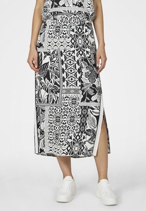 Frau trägt einen schwarz-weiß gemusterten Midirock mit Seitenschlitz und weißen Sneakers, steht vor einem schlichten Hintergrund.