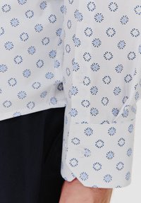 Biała koszula z niebieskim okrągłym wzorem, z zapięciem na guziki i usztywnionym kołnierzykiem. Długi rękaw z elegancko dopasowanym mankietem.