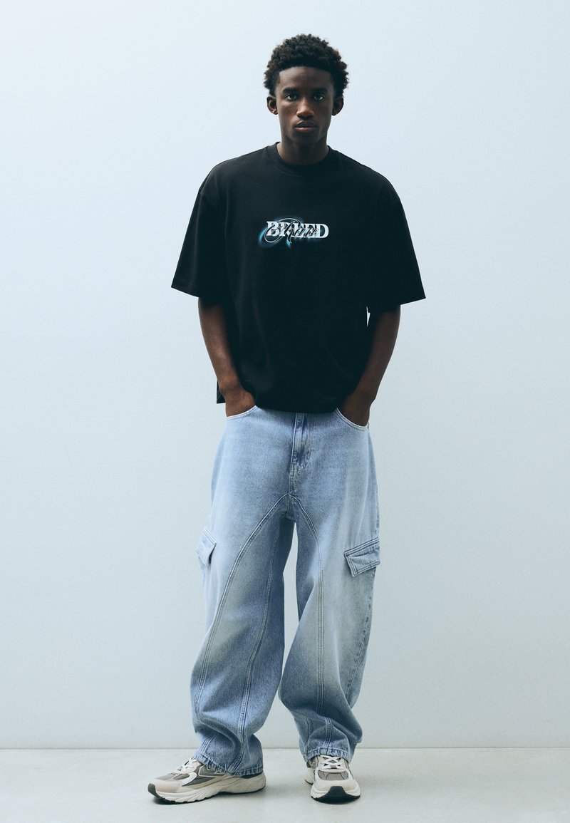 Jeune homme aux cheveux courts et bouclés portant un t-shirt noir à motif, un jean cargo ample bleu clair et des baskets blanches, debout contre un mur uni.