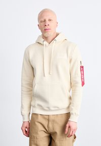 Haut à capuche beige avec cordons de serrage, poche avant et un écusson sur la poitrine. Présente une étiquette rouge sur la manche gauche avec du texte.