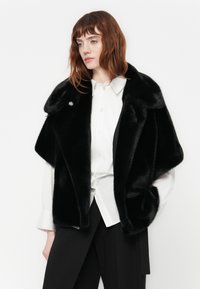 Zwarte faux fur jas met brede revers, voorzien van een enkele decoratieve knop en een zachte, pluche textuur. Draag over een wit shirt en zwarte broek.