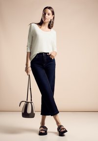 Hellgrünes, langärmliges Oberteil kombiniert mit dunkelblauen, weit geschnittenen Jeans. Das Model hält eine graue und cremefarbene Bucket Bag und trägt schwarze Sandalen mit Schnallen.