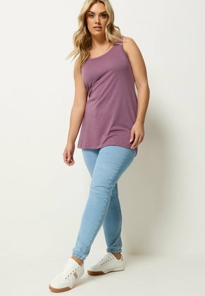 Vaaleahiuksinen nainen, jolla on päällään hihaton violetti paita, vaaleansiniset kapealahkeiset farkut ja valkoiset tennarit, seisoo yksivärisen taustan edessä.