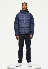 Mørkeblå pufferjakke med vandret syninger, fast hætte, lynlås lukning, parret med mørke jeans og hvide sneakers, stående på en cirkulær pedestal.