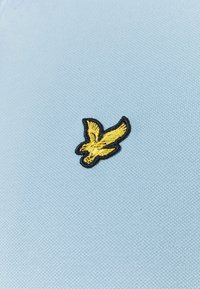 Polo shirt azzurro chiaro con logo di un uccello giallo, ricamato con contorni neri, su un tessuto testurizzato.