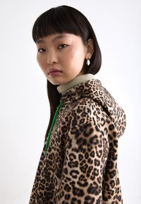 Luipaardprint hoodie met een zachte textuur, groene trekkoorden en een beige basis, gecombineerd met een hoogsluitend top in een subtiele beige kleur.