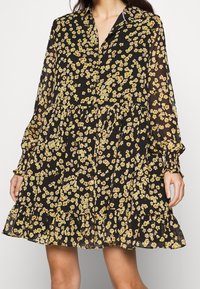 Zwarte bloementop met levendige gele en rode bloemen, lange mouwen, knoopsluiting en een gelaagde rok met een gerimpelde zoom. Lichtgewicht stof.