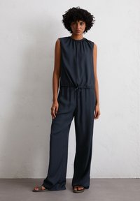 Frau mit lockigem Haar, die einen ärmellosen, dunkelblauen Jumpsuit mit Kordelzug und weit geschnittenen Hosen trägt und vor einer strukturierten weißen Wand steht.