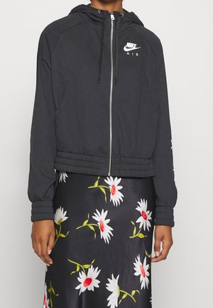 Person iført en sort Nike Air lynlås-hættetrøje med hætte og et sort skørt med et hvidt og rødt margueritmønster.