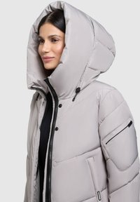 Chaqueta acolchada gris claro con una gran capucha, cierre de cremallera y botones de presión, que cuenta con un bolsillo lateral con cremallera y una etiqueta de la marca en el dobladillo.