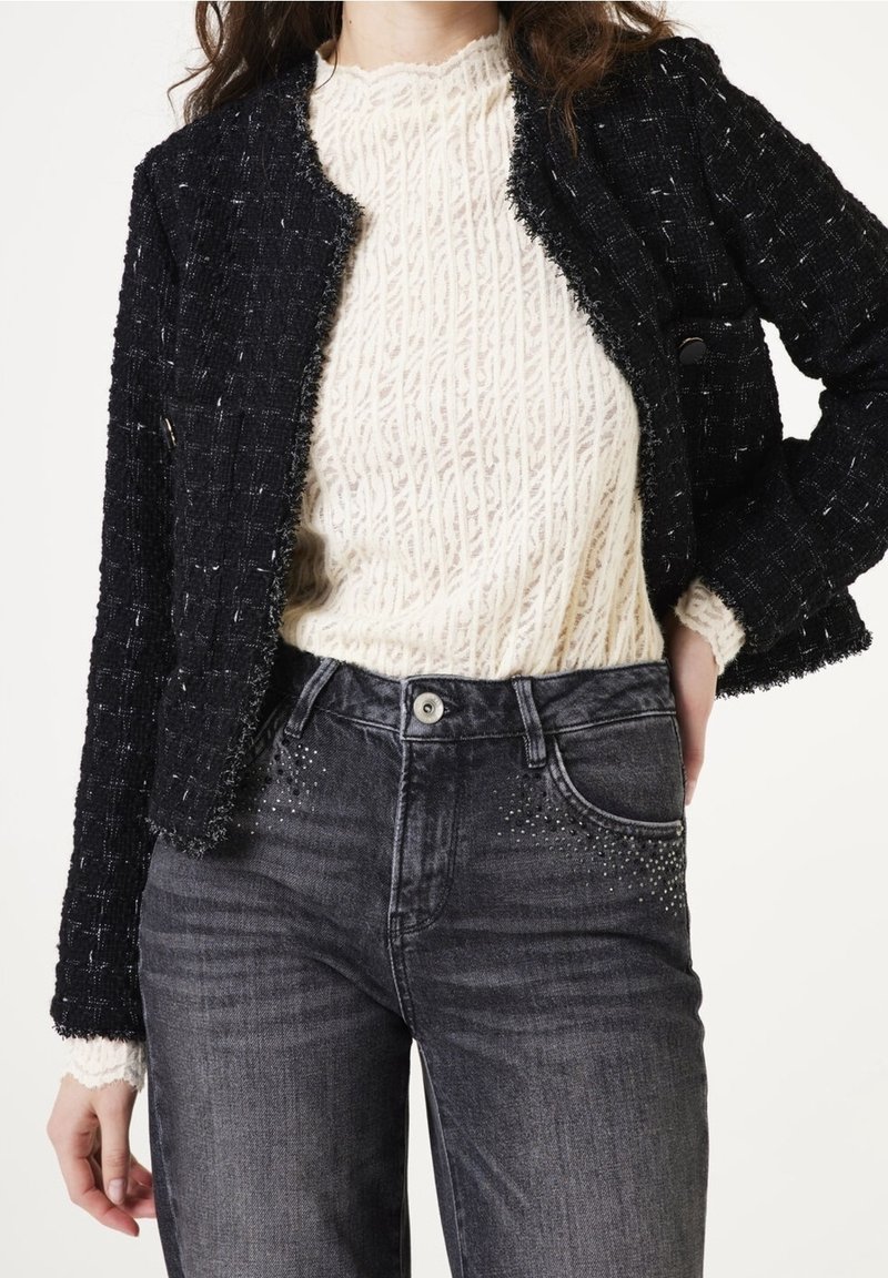 Femme portant un haut en dentelle crème à manches longues, une veste noire texturée ouverte et un jean gris foncé taille haute avec de petites décorations sur les poches.