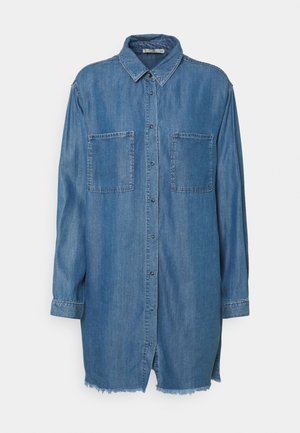 Vestido camisa de mezclilla azul de manga larga con cuello, botones en el frente, dos bolsillos en el pecho y dobladillo deshilachado sobre fondo blanco.