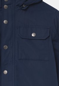 Giacca blu navy realizzata in materiale resistente all'acqua, con chiusura a zip, bottoni a pressione e tasca sul petto con pattella e bottone a pressione.