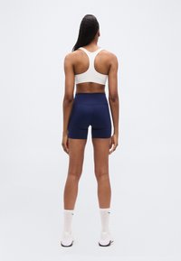 Λευκό αθλητικό σουτιέν με σχέδιο racerback, συνδυασμένο με σορτς σε ναυτικό μπλε. Η στολή είναι κατασκευασμένη από ελαστικό ύφασμα, κατάλληλη για αθλητικές δραστηριότητες.