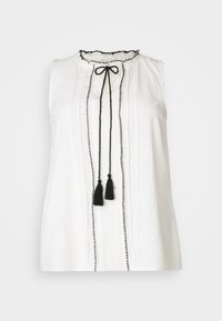 Bílý bezramínkový top z lehkého materiálu s volánkovým výstřihem, černým lemem, centrálním skladem a dvěma černými třásněmi vpředu.