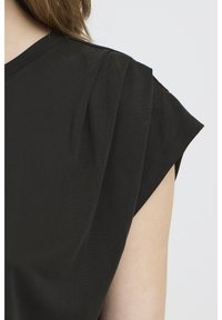 Približni posnetek črnega kratkoročnega vrha z nežnimi naborki na ramenih na osebi s svetlo poltjo in rjavimi lasmi.