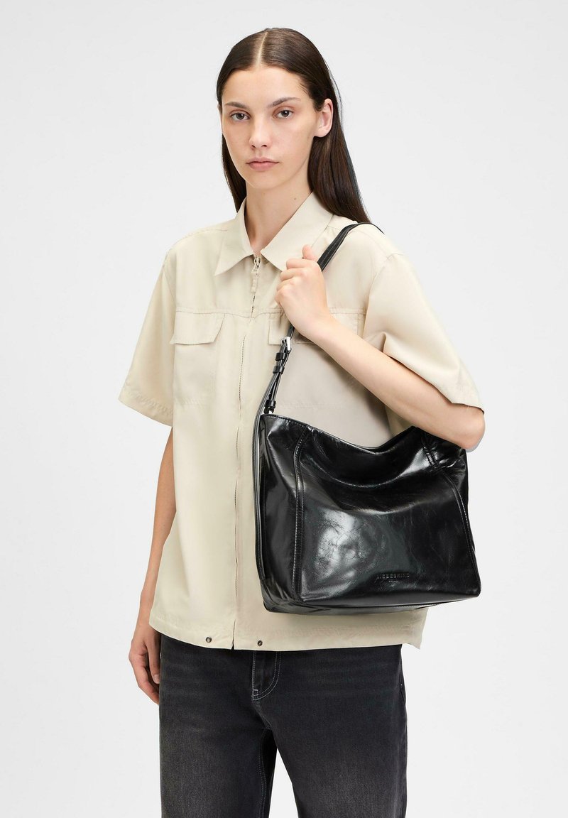 Schwarze Leder-Shoulderbag mit glänzender Oberfläche, kurvenförmigem Design und verstellbarem Tragegurt, präsentiert von einer Person in einem hellbeigen Hemd.