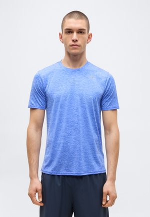 Młody mężczyzna z bardzo krótkimi włosami, ubrany w niebieską sportową koszulkę z krótkim rękawem i ciemne spodenki, stoi na jednolitym jasnym tle.